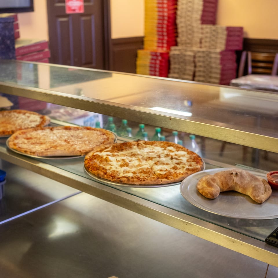 Three cheese pizzas and a calzone with sauce on a display shelf in a pizzeria.
