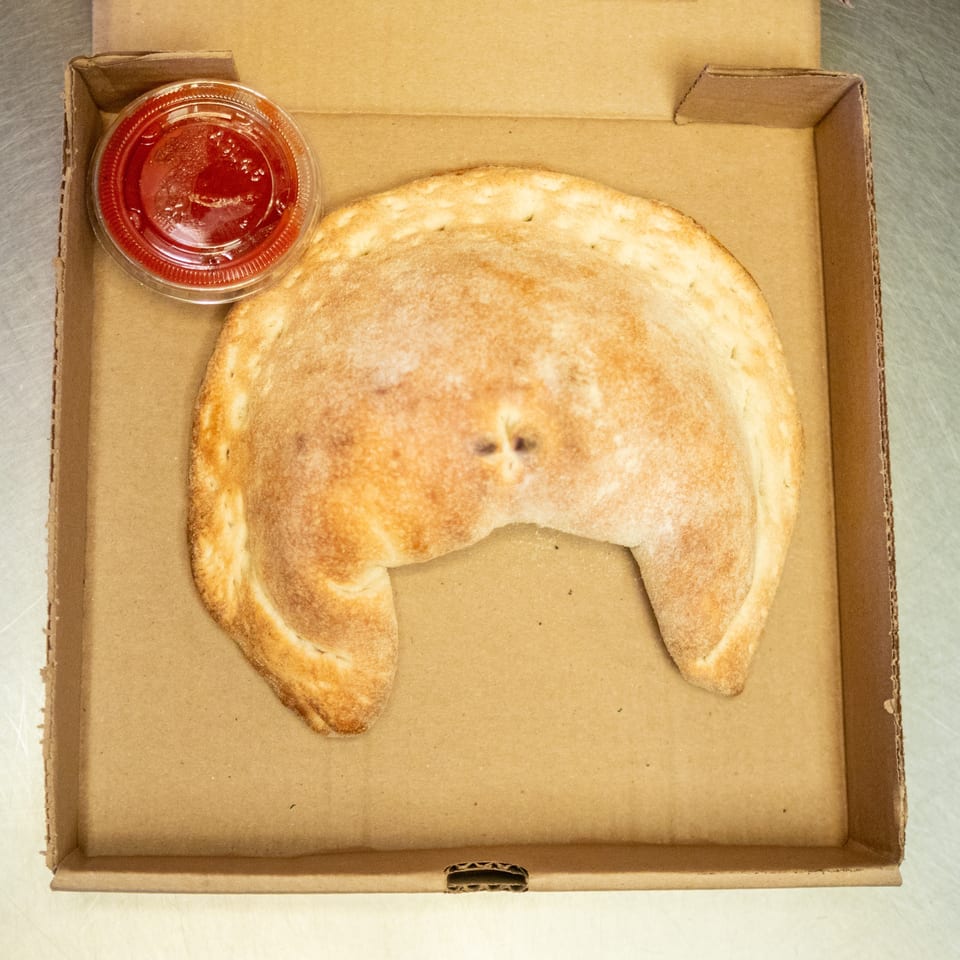 A golden-brown calzone and a small cup of red sauce in a cardboard pizza box.