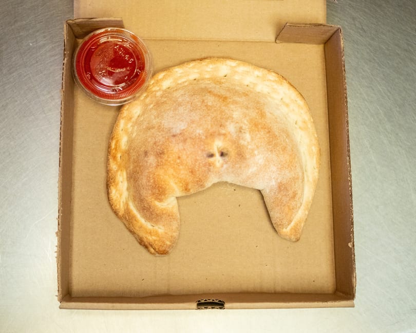A golden-brown calzone and a small cup of red sauce in a cardboard pizza box.