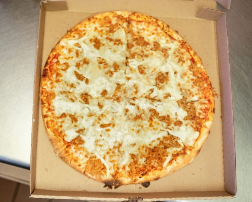 A whole, round pizza with melted cheese and sausage crumbles sits in a cardboard delivery box.