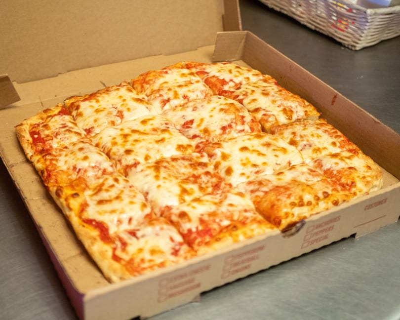 A square cheese pizza, cut into many small squares, sits in an open cardboard box.