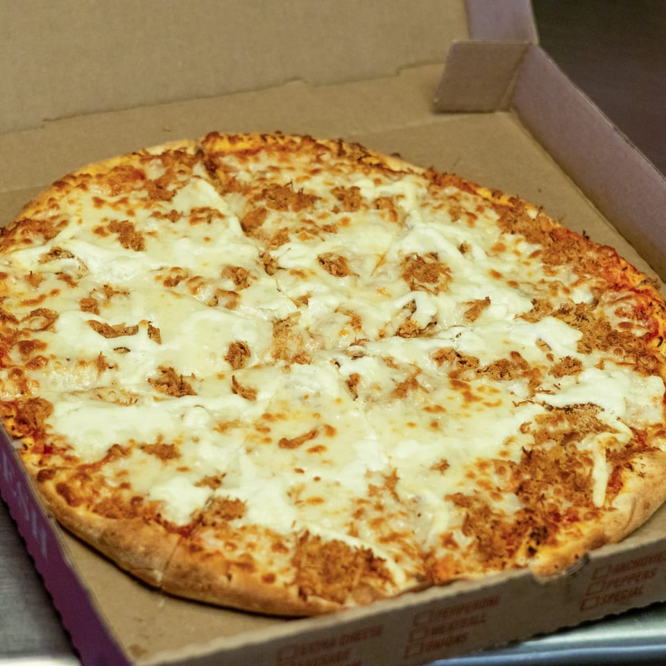 A whole pizza with white cheese and shredded meat toppings in a cardboard box.