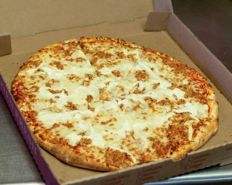 A whole pizza with white cheese and shredded meat toppings in a cardboard box.