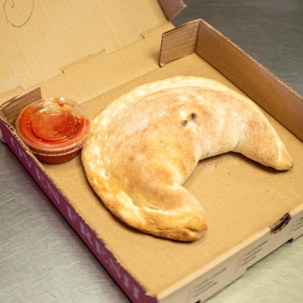 A golden-brown calzone and a small cup of red sauce sit inside an open cardboard box.