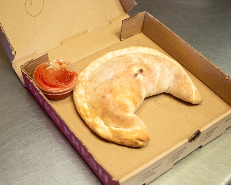 A golden-brown calzone and a small cup of red sauce sit inside an open cardboard box.